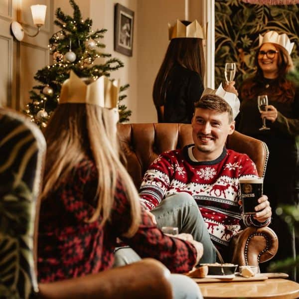 Two colleagues in christmas wear sitting in cosy seats at a pub. 2 others are in the background. The theme is christmasy